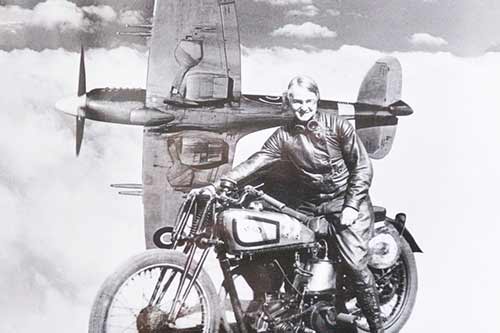 Old black and white photo of a lady sat on a motorbike with a backdrop of clouds and mini aeroplane on its side