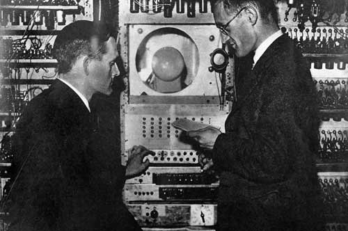 Old black and white photo of two men operating a large metal machine that has lots of buttons and wire on it