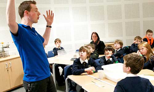 Male teacher explaining something to a class of children with his hands high in the air