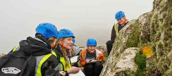 Four students with clipboards and wearing hard helmets looking at a large moss-covered rock