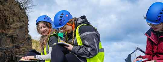 Students holding clipboards wearing hard hats and sat on a rock