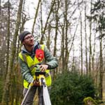 Man stood holding a metal tripod that supports a lens, outside beside tall trees and wearing a high-visibility jacket.